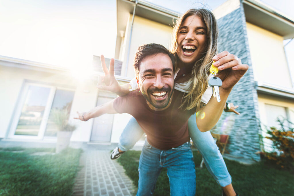 Happy young couple holding home keys after buying real estate – Husband and wife standing outside in front of their new house
